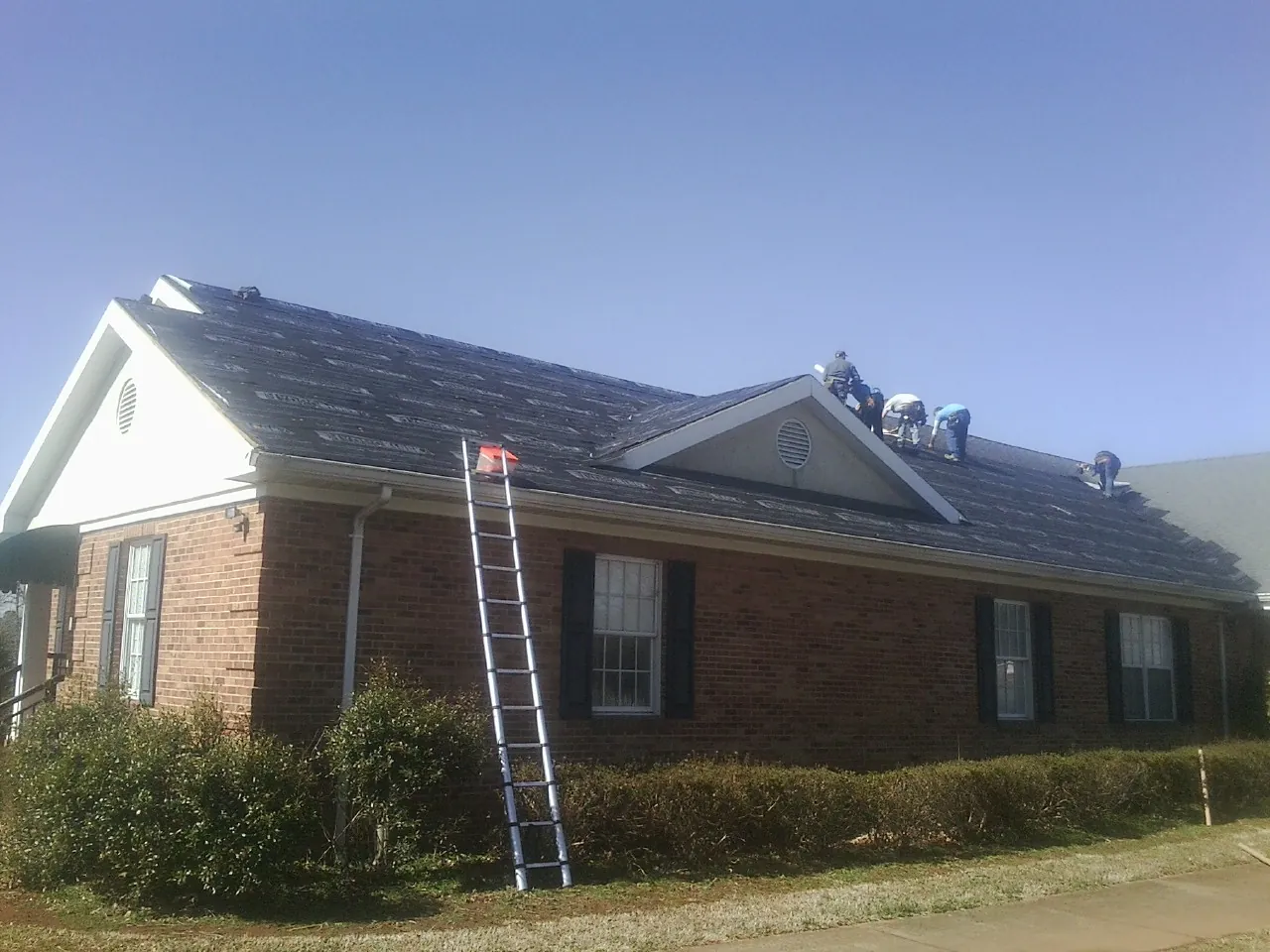 Crew tearing shingles off a brick ranch roof with a ladder set — Metal Roof Repair work by our crew in Monticello